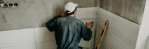 A man in blue work coveralls kneels in a bathroom corner to install large white tiles on the wall.